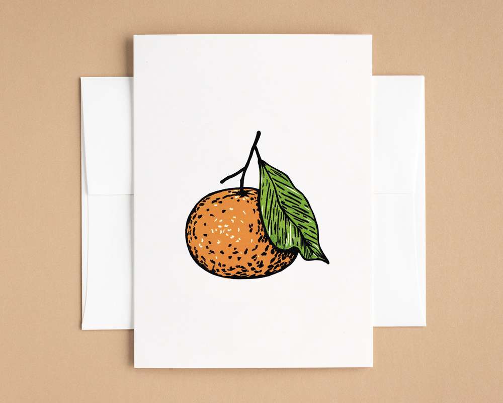 A vertical white card depicts a mandarin orange using bold black lines filled in with orange and green. The card sits on top of a white envelope, which lies on a brown backdrop.